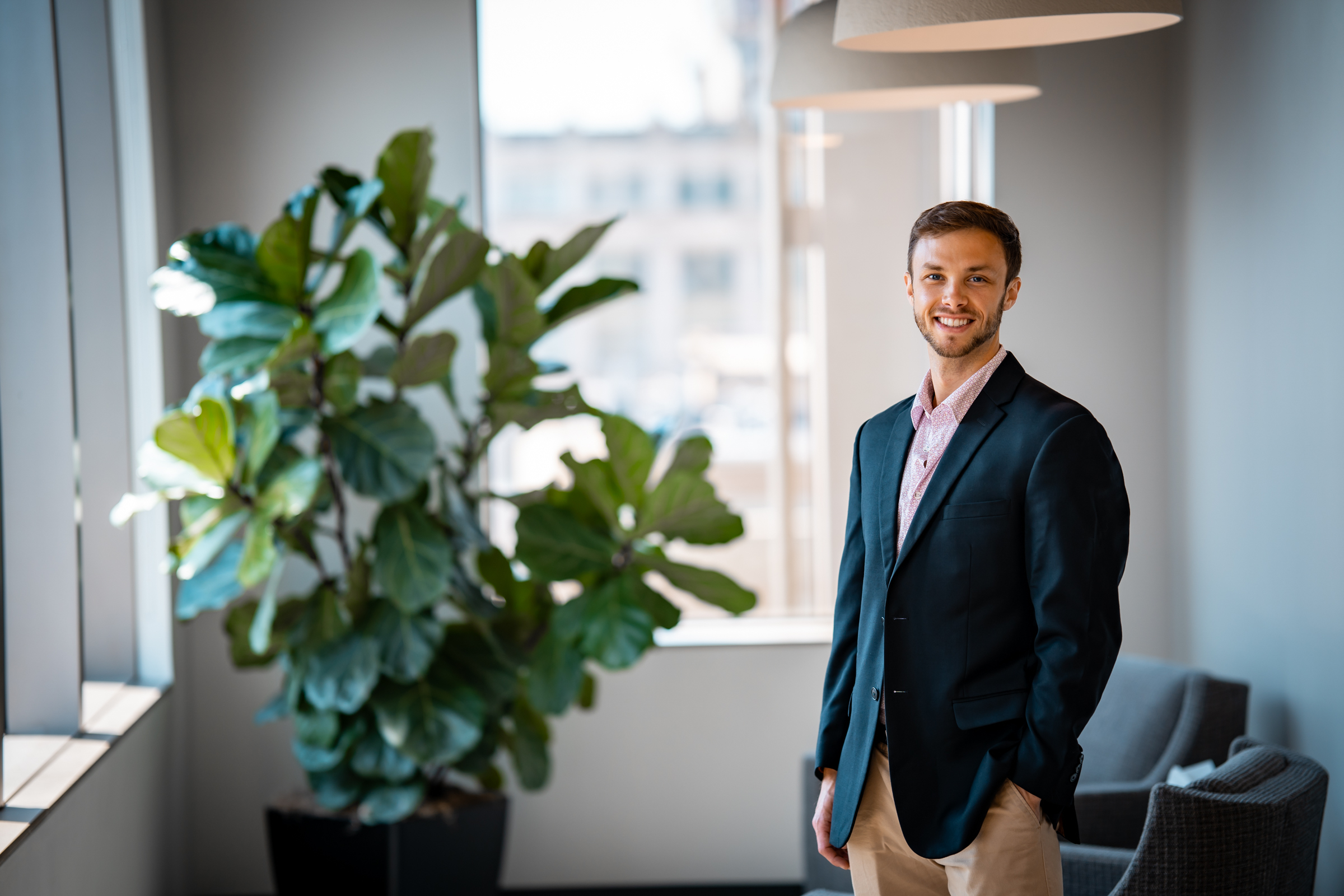 Photo of Keenan Hawekotte standing in the office and smiling next to a fiddle leaf fig tree