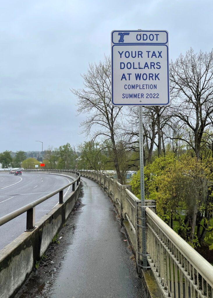 Image of a sign near a road and sidewalk that says "ODOT Your tax dollars at work completion summer 2022"